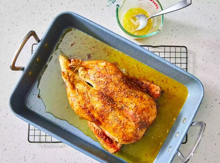 A roasted duck resting in a roasting pan, with a small cup of melted butter beside it