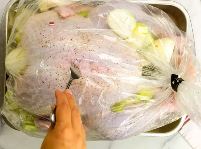 A hand using a fork to create holes in a sealed turkey bag, with raw turkey and vegetables inside.