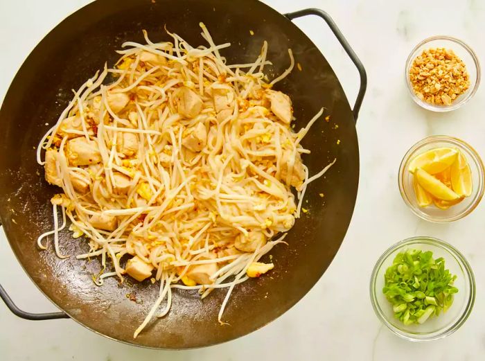 Aerial shot of chicken, eggs, noodles, and bean sprouts cooking together in a wok, with other pad Thai ingredients in bowls to the side.