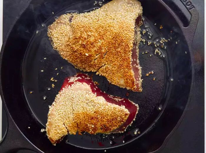 A cast iron skillet with two tuna steaks seared with sesame crust.