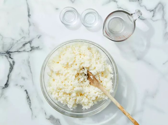 The cooked rice is placed in a bowl, and vinegar, salt, and sugar are mixed in.