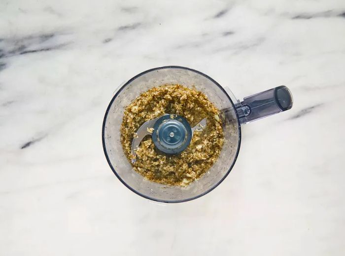 A top-down view of garlic, onion, mint, salt, and pepper mixed together in a food processor.