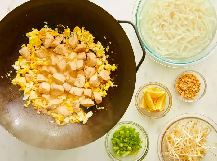 Aerial view of chicken and eggs cooking in a wok, with noodles and other pad Thai ingredients placed in bowls nearby.