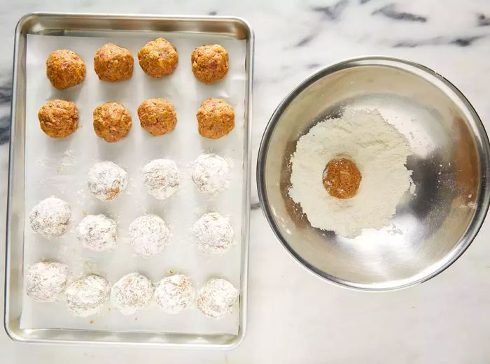 A top-down view of meatballs being coated in flour and arranged on a baking sheet.