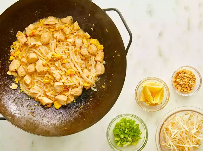 Aerial view of chicken, eggs, and noodles cooking in a wok, with additional pad Thai ingredients in bowls nearby.