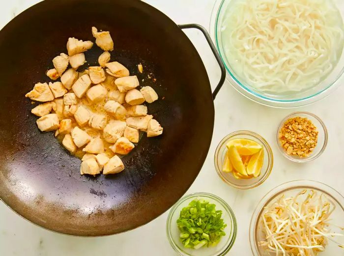 Bird's-eye view of chicken being cooked in a wok, surrounded by noodles and other pad Thai ingredients in separate bowls nearby.