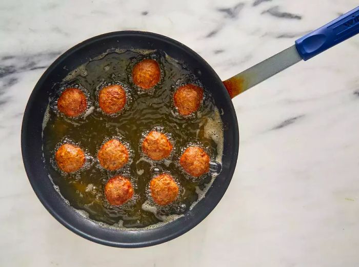 A top-down view of meatballs frying in oil in a skillet.