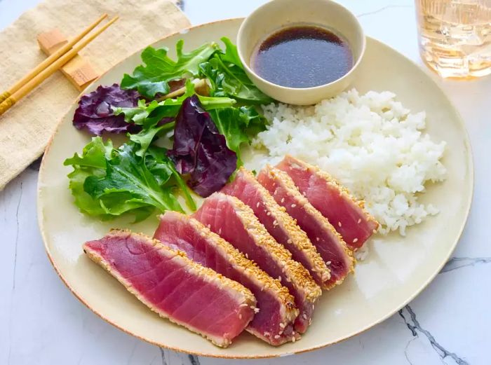 A plate of sliced sesame-seared tuna served with a sauce, accompanied by a green salad and white rice.