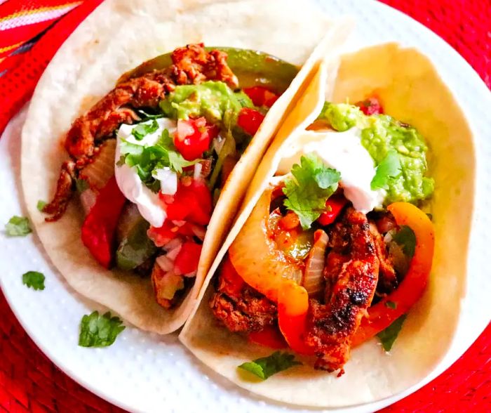 A top-down shot of two flour tortillas filled with a vibrant blend of grilled chicken breast strips, sautéed yellow and red bell peppers, onions, and topped with a dollop of sour cream, guacamole, and fresh cilantro.