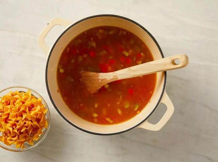 A top-down view of carrots, onions, celery, thyme, salt, and broth cooking in a pot, with a side of noodles nearby.