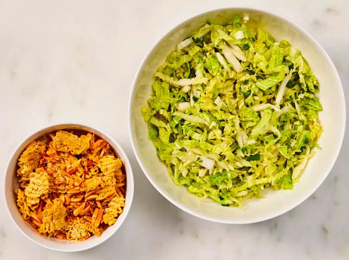 Two bowls: one filled with chopped cabbage, the other with crispy toasted ramen and almonds.