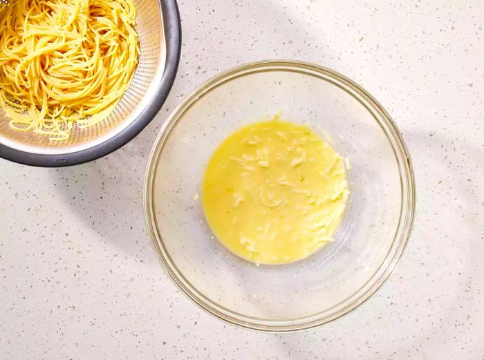 Spaghetti draining in a colander beside a glass bowl filled with the egg and cheese mixture