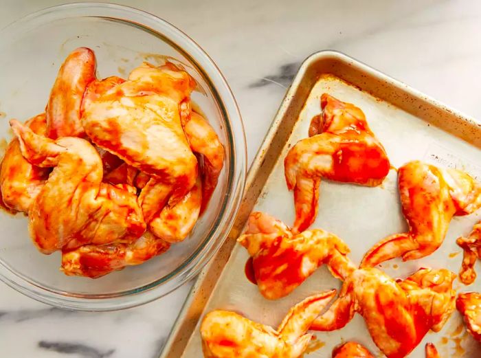 Top-down view of raw chicken wings coated in sauce, with some in a bowl and others on a baking sheet.