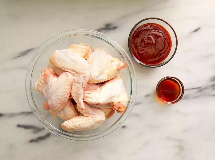 View from above of raw chicken wings in a bowl, with sauces placed beside them.