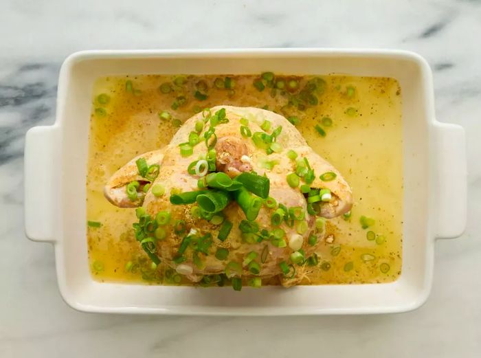 A baking dish with a whole chicken sitting upright on a beer can, seasoned with garlic mix and topped with sliced green onions