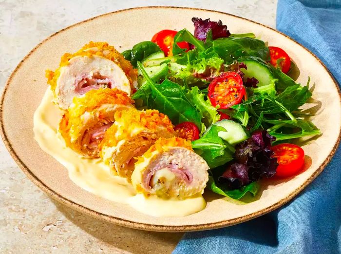 A sliced chicken cordon bleu roll served on a plate, with a side of cordon bleu sauce and a fresh mixed greens salad.