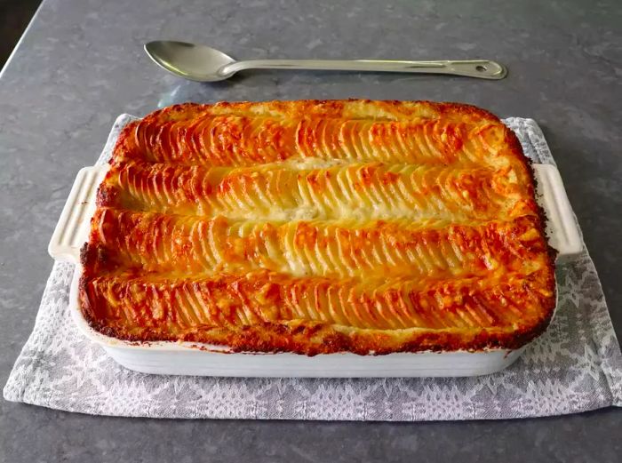 A golden, crispy casserole of potatoes au gratin resting on a grey placemat with a spoon beside it.