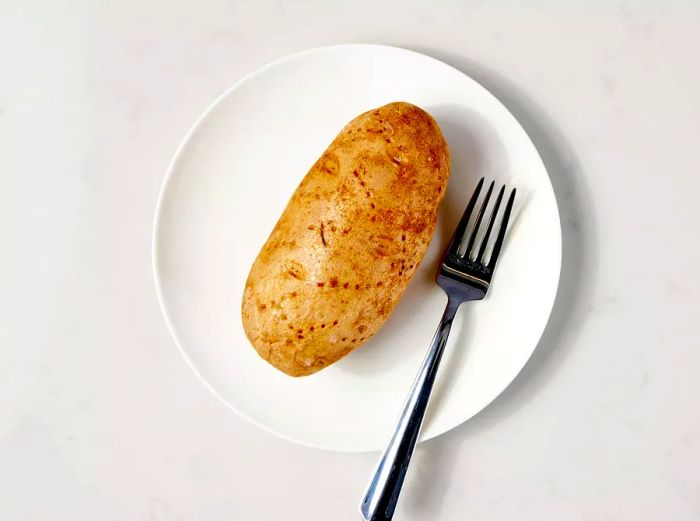 A raw russet potato resting on a white plate with a fork beside it.