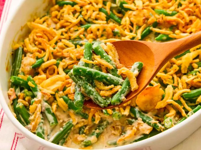 A wooden spoon serving a portion of Campbell's green bean casserole, placed beside the casserole dish