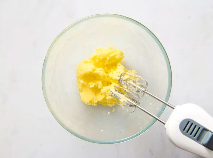 A large glass mixing bowl with sugar and butter being blended using a hand mixer