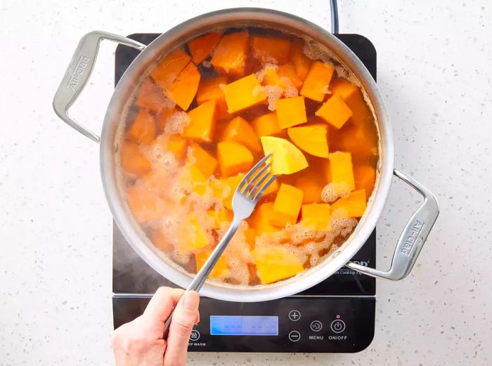 A pot with peeled sweet potato chunks in boiling water, with a fork inserted into a piece of sweet potato to check for tenderness