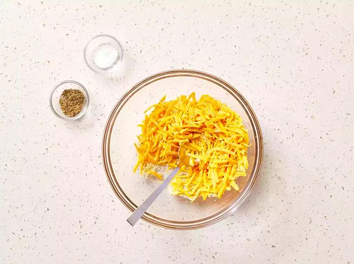 A large bowl filled with Cheddar, Parmesan, garlic, oregano, and basil, accompanied by small bowls of salt and pepper.