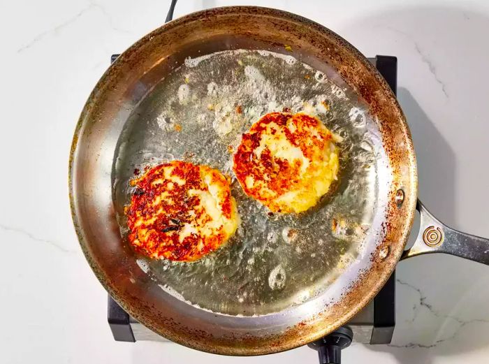 A skillet with two golden-brown potato cakes frying in hot oil.