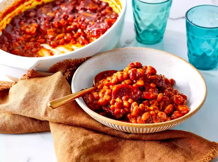 A large bowl of baked beans with extra beans in a white baking dish in the background.