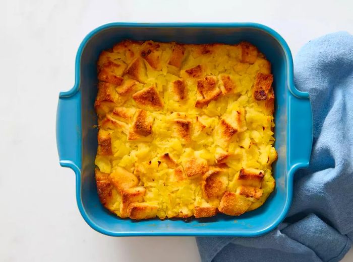 A square baking dish filled with golden, baked pineapple stuffing, with a blue kitchen towel nearby