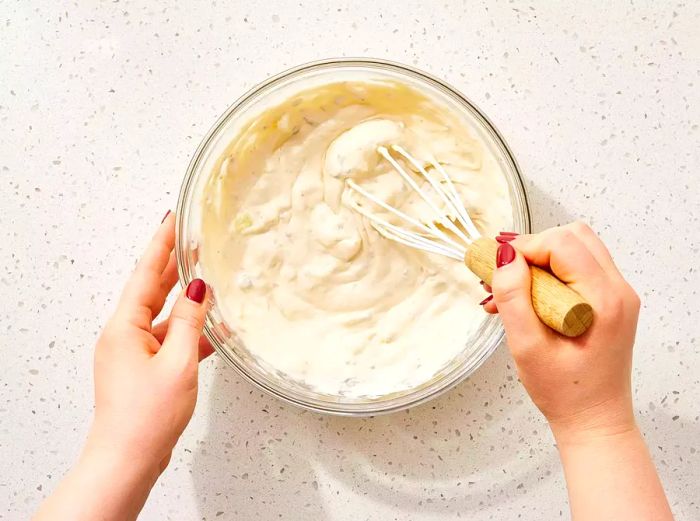 A bowl filled with whisked condensed milk, mayonnaise, egg, and onion mixture.