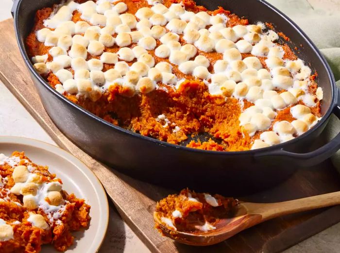 A baking dish filled with sweet potato casserole, topped with golden roasted marshmallows, alongside a plated serving.