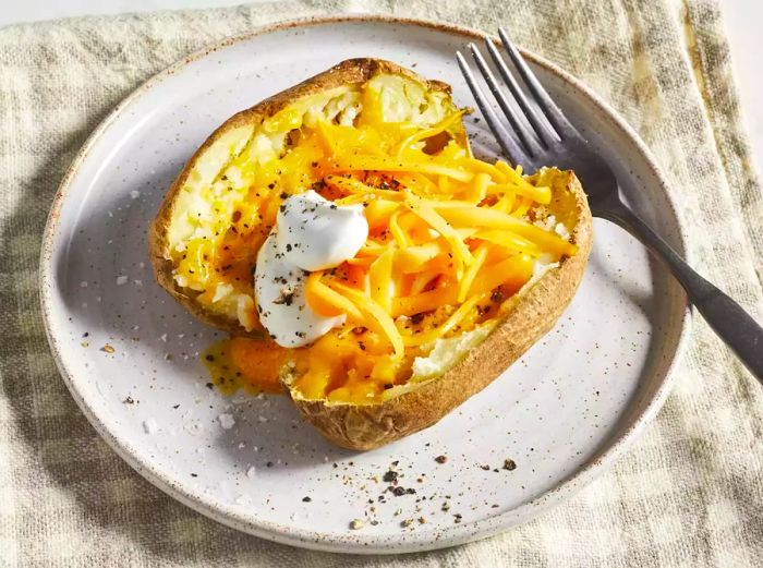 A close-up view of a perfectly cooked microwave baked potato topped with melted cheese, sour cream, salt, and pepper, served on a plate with a fork beside it.