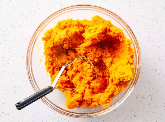 A glass bowl filled with mashed sweet potatoes, accompanied by a potato masher