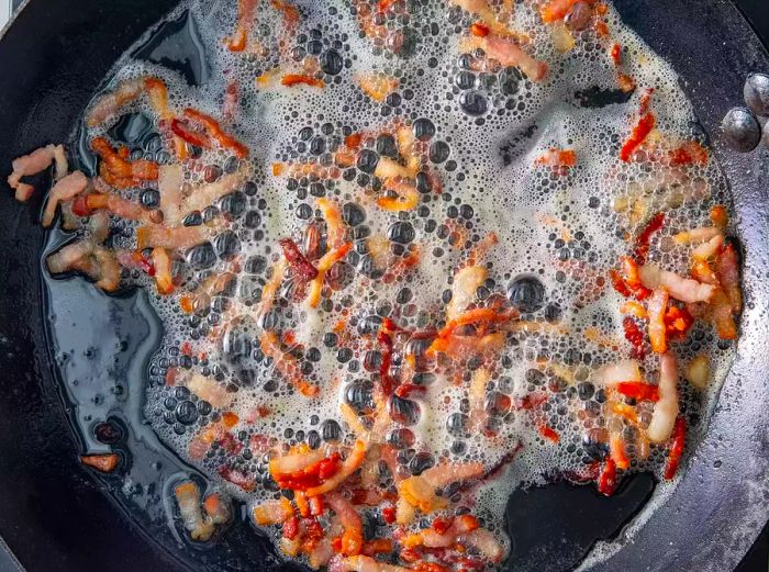 A bird’s-eye view of bacon pieces sizzling in a cast-iron skillet.