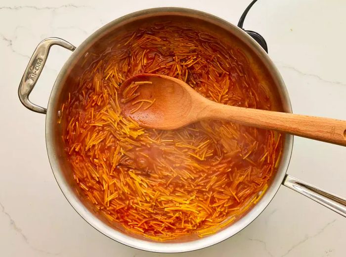 A pot of fideo pasta simmering in seasoned tomato sauce and water, stirred with a wooden spoon