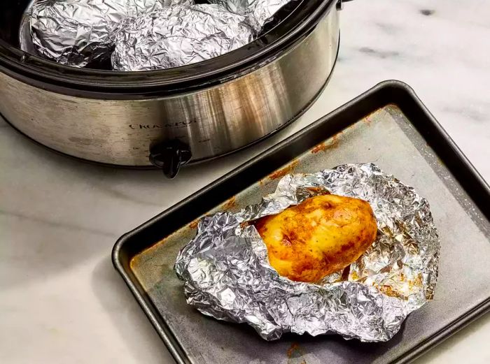 A slow cooker filled with foil-wrapped potatoes, next to a baking sheet with a baked potato also wrapped in foil.