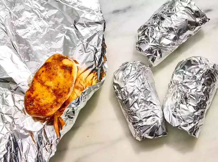 A potato coated with oil and wrapped in foil, placed next to three other foil-wrapped potatoes.