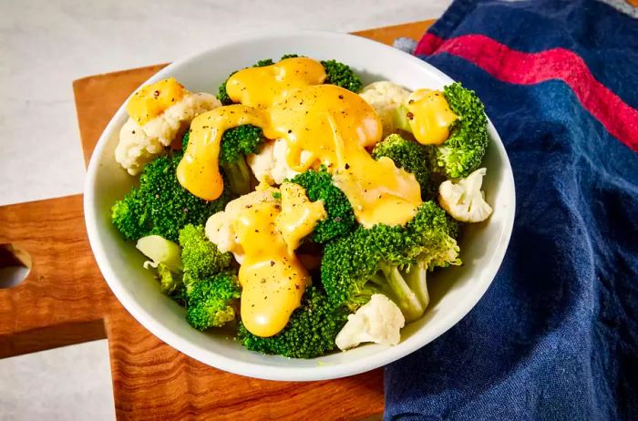 A plate of steamed broccoli and cauliflower drizzled with cheese sauce and sprinkled with ground black pepper, served on a wooden board with a kitchen towel.