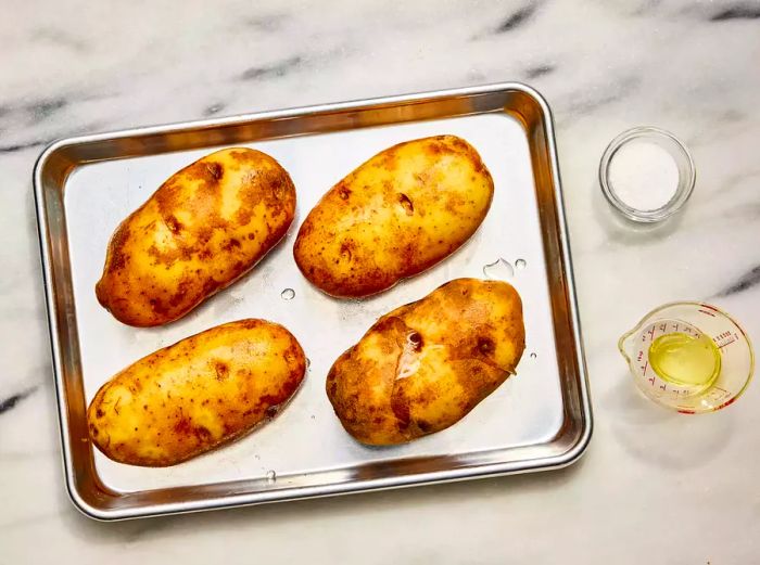 A baking sheet with four whole potatoes, coated in oil and sprinkled with salt.