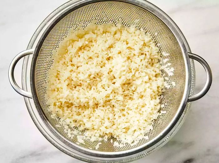 Overhead view of sushi rice being rinsed in a strainer.