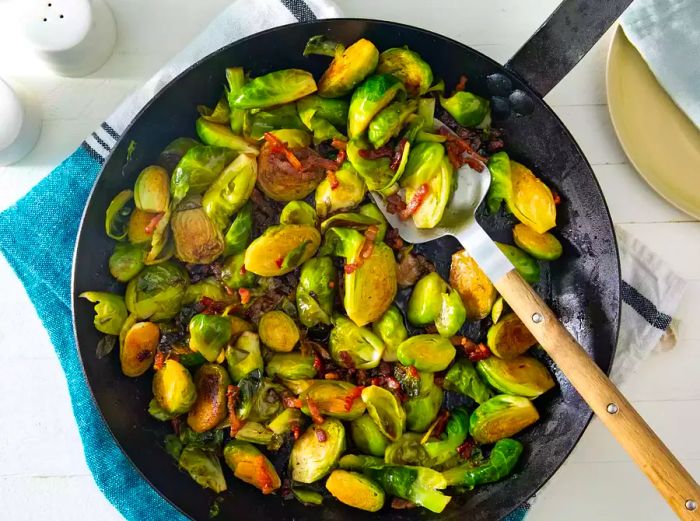 An overhead shot of a black skillet filled with Brussels sprouts, bacon, and a spoon.