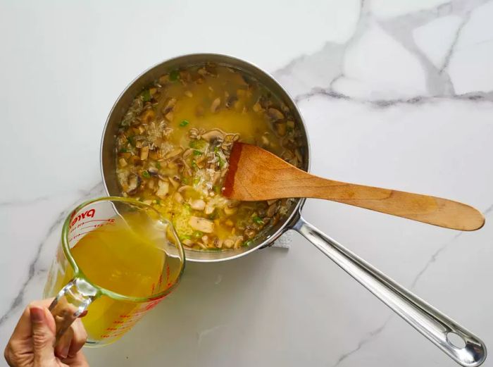 A top-down view showing chicken broth being poured into a pot of mushroom rice, with a wooden spoon stirring.