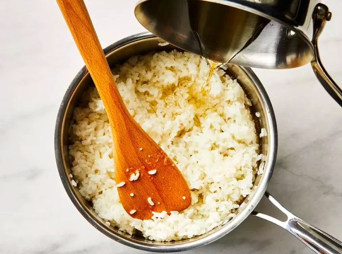 Vinegar mixture being poured into a pot with sushi rice, stirred with a wooden spatula.