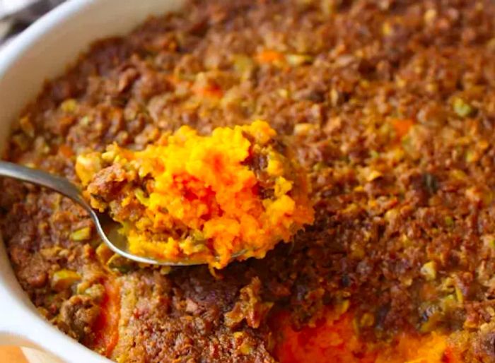 Close-up shot of a spoon lifting a serving of pistachio-crusted sweet potato casserole from a white oval baking dish.