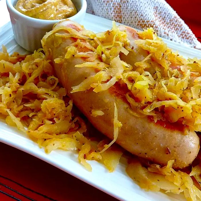 Two bratwurst sausages topped with sauerkraut, served with a side of mustard in a small ramekin on a square ceramic plate