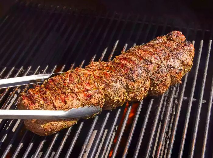 Tongs holding a beef tenderloin with herb-garlic-pepper coating on the grill.