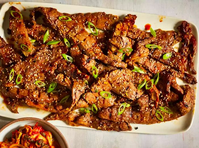 A close-up overhead shot of a platter of savory beef bulgogi, garnished with sesame seeds and sliced green onions, served alongside kimchi.