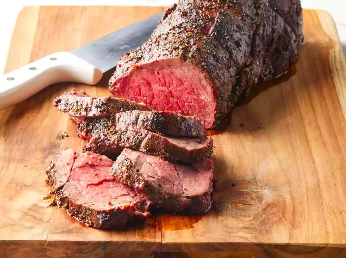 Sliced grilled beef tenderloin with herb-garlic-pepper coating, resting on a wooden cutting board next to a knife.