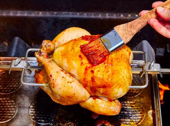 A whole chicken on a rotisserie attachment over a grill, being brushed with seasoned melted butter