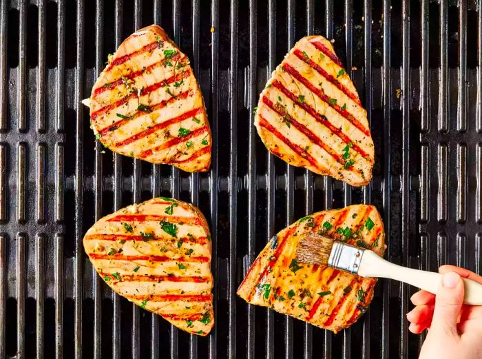 Four marinated tuna steaks being basted while grilling.